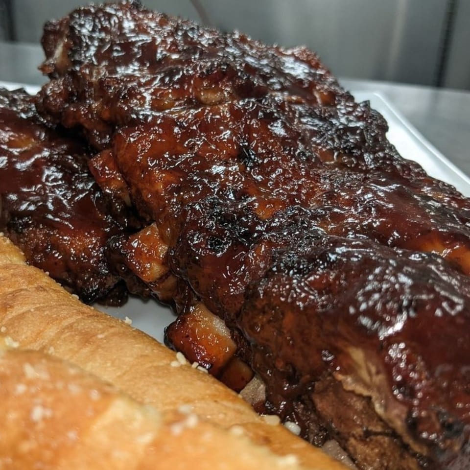 Close-up of glistening BBQ ribs with a side of golden-brown bread from Hippo's Chicken, Pizza, Ribs & Fish.
