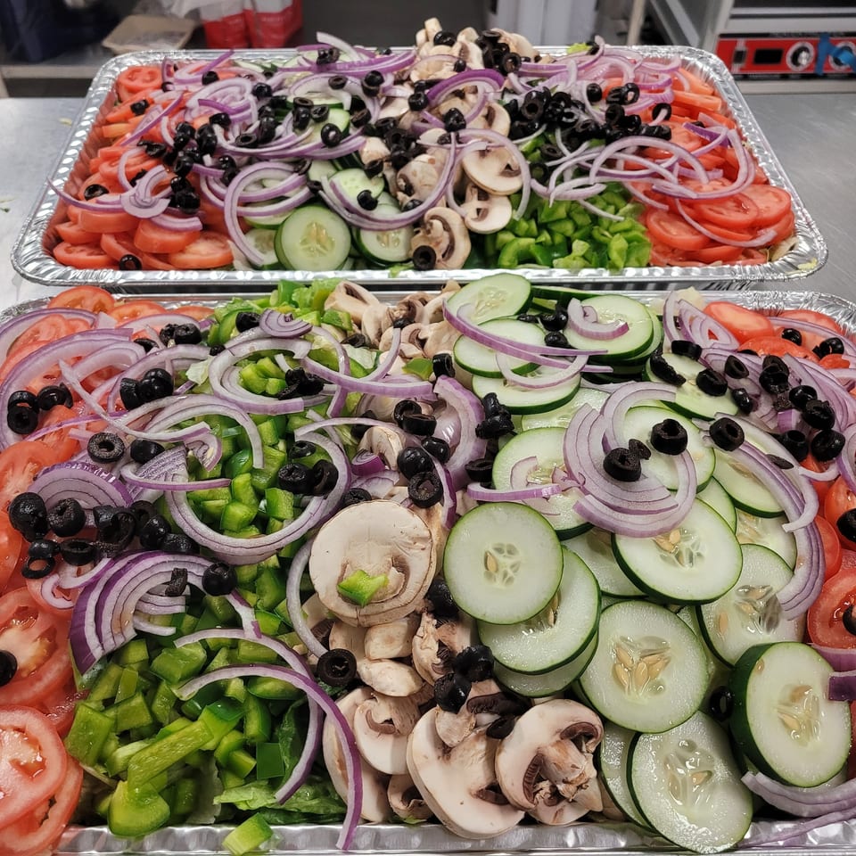 Two large foil trays filled with fresh salad toppings: tomatoes, onions, cucumbers, mushrooms, and olives.