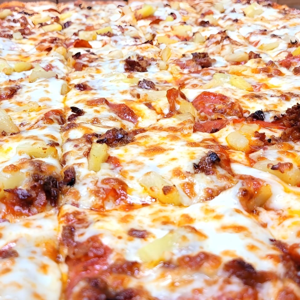 Close-up of a Hippo's pizza with melted cheese, pineapple, and meat toppings, cut into squares.