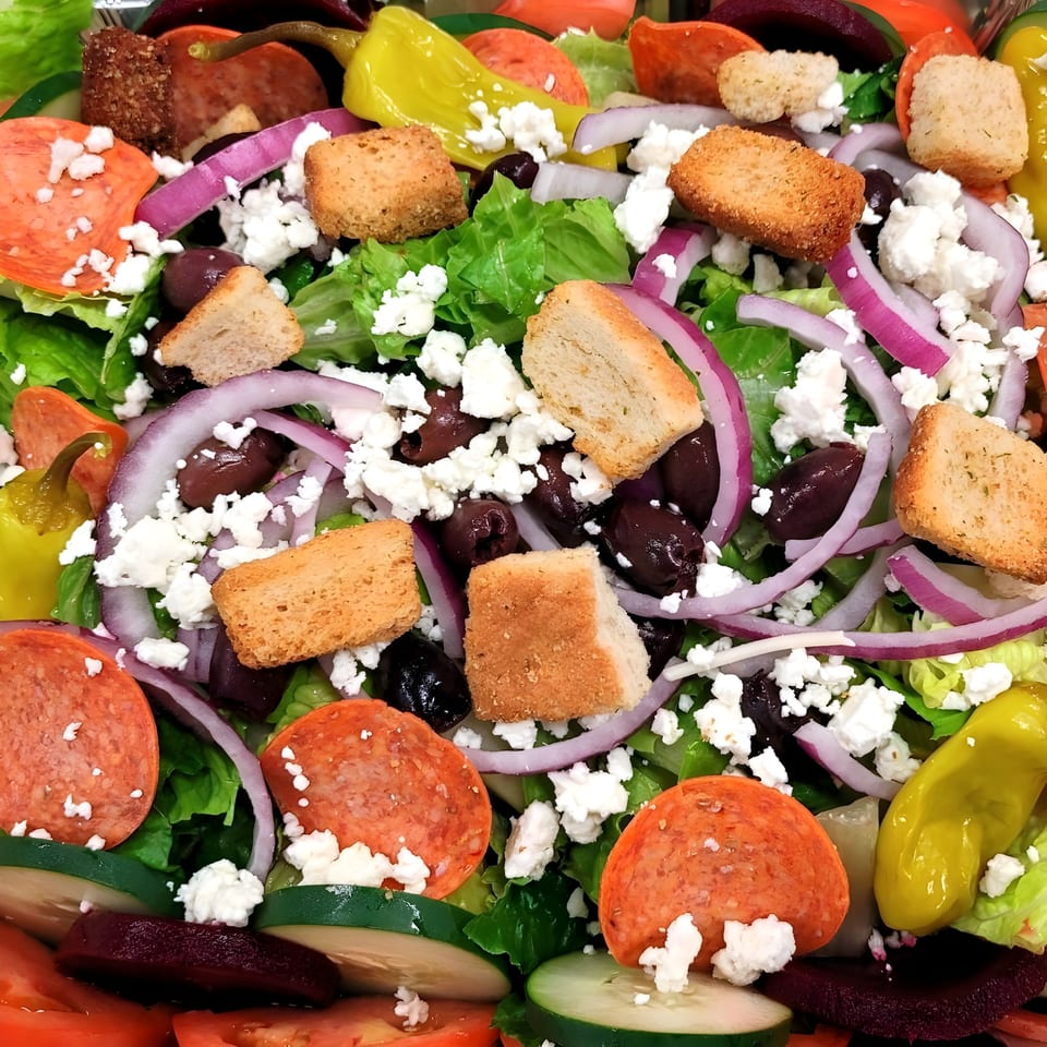 Close-up of a vibrant salad with pepperoni, feta, olives, red onion, croutons, and lettuce.